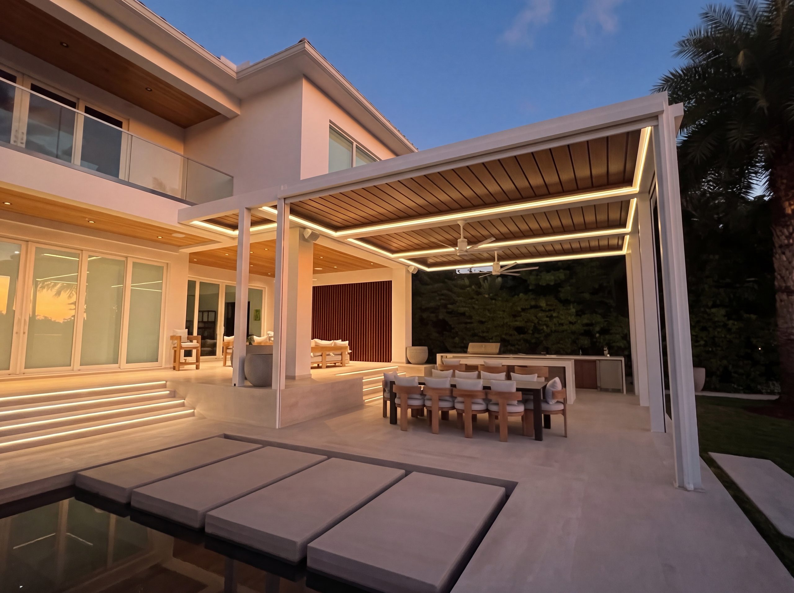 A Decopatio bioclimatic pergola at twilight — lit louvered roof over a terrace dining lounge, beside the pool